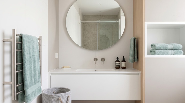 Colores claros para hacer que un baño pequeño parezca más amplio Baño pequeño moderno en tonos blancos con mueble suspendido, espejo redondo y toallas verde agua que aportan mayor sensación de amplitud.